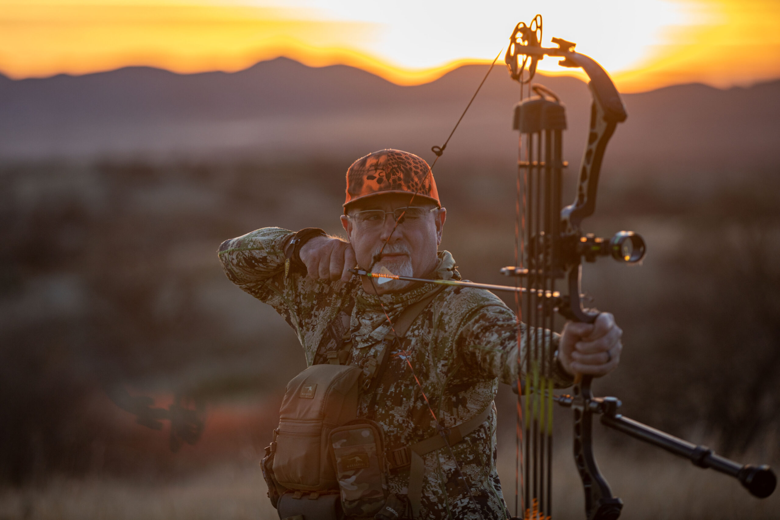 A man in camouflage holding a bow and arrow at home.